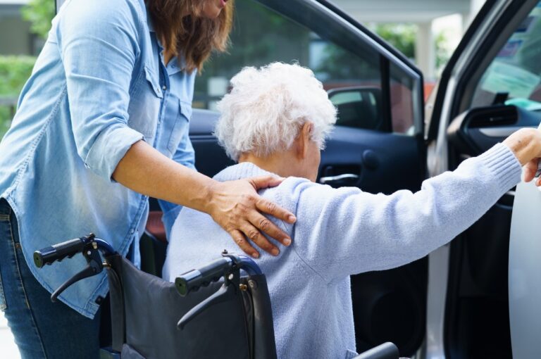 Pflegerin hilft asiatischer älterer Frau mit Behinderung im Rollstuhl, ins Auto zu steigen.